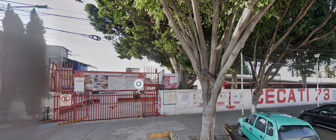 Fotografía en la que se muestra la entrada del plantel, conformada por una reja de color rojo a través de la cual se puede ingresar a las instalaciones. A la derecha de la reja hay un muro blanco que tiene escrito en letras grandes de color rojo CECATI 73.